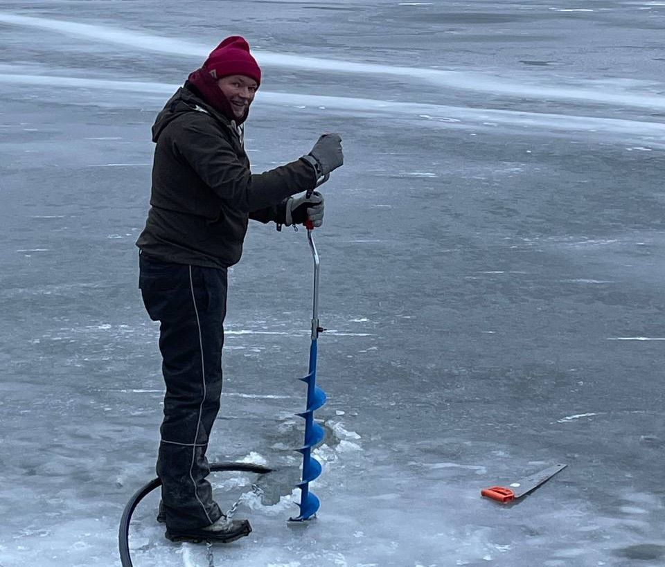 Per Axbom standing on a frozen lake, using a manual drill to bore a hole. 