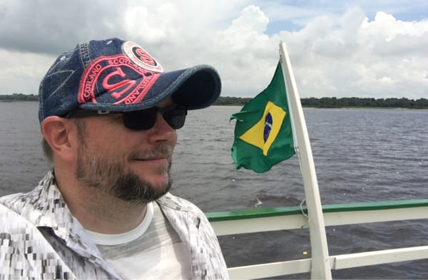 Closeup of Per Axbom, wearing a baseball hat and sunglasses, on a boat with water in the background and land in the distance.