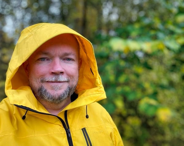 Close-up of smiling Per Axbom in a yellow raincoat with the hood up. Greenery in the background.